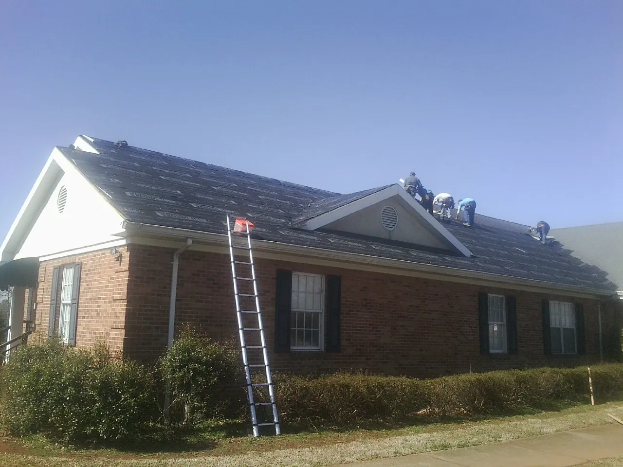 Crew tearing shingles off a brick ranch roof with a ladder set — Emergency Roof Repair work by our crew in Pine Brook
