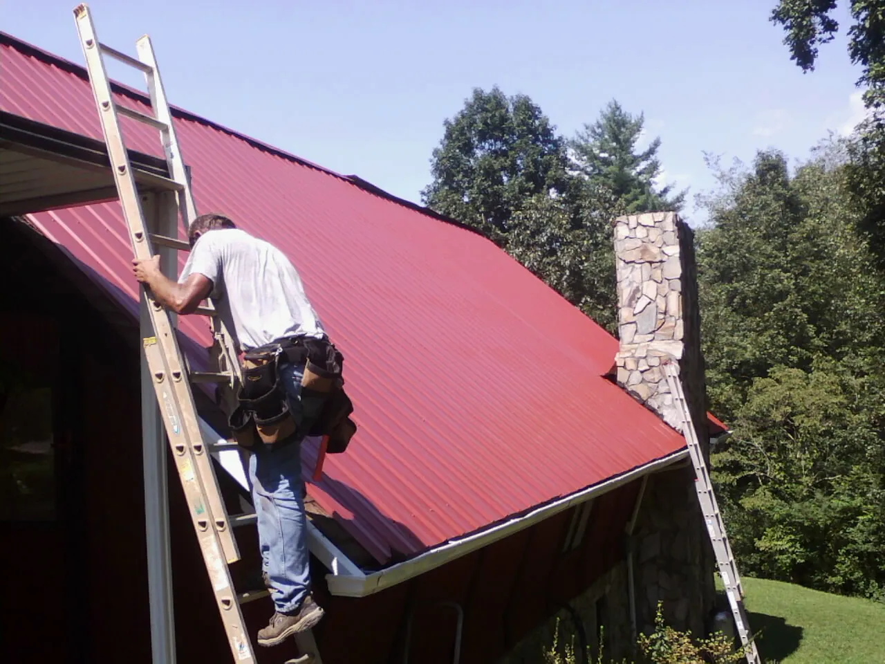 Roofer on a ladder beside a red metal roof and stone chimney — Soffit & Fascia Repair by Roofing Brothers in Pine Brook